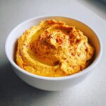 A close-up of creamy, vibrant orange Roasted Red Pepper Hummus swirled with olive oil and sprinkled with paprika in a white bowl.