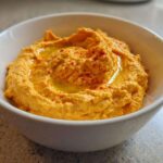 Close-up of creamy Roasted Red Pepper Hummus drizzled with olive oil and sprinkled with paprika in a white bowl.