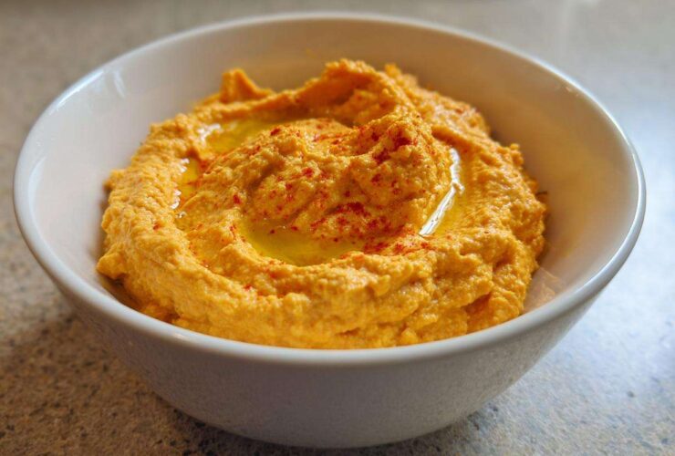 Close-up of creamy Roasted Red Pepper Hummus drizzled with olive oil and sprinkled with paprika in a white bowl.