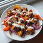 A white bowl filled with Sheet Pan Chicken Shawarma pieces, roasted peppers, and red onions, drizzled generously with garlic yogurt sauce.