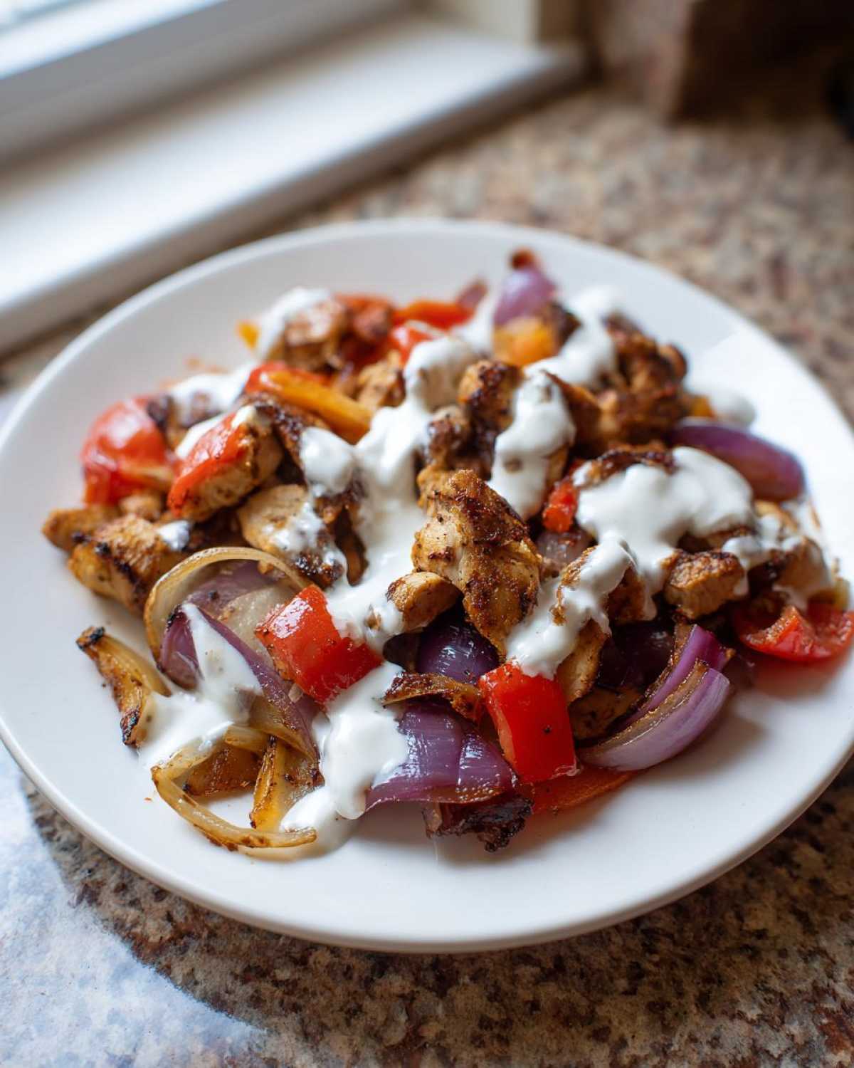 Close-up of Sheet Pan Chicken Shawarma pieces mixed with roasted red peppers and onions, generously drizzled with garlic yogurt sauce.