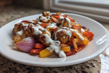 A serving of Sheet Pan Chicken Shawarma with roasted peppers and onions, drizzled generously with garlic yogurt sauce.