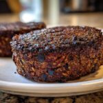 Close-up of a thick, richly seasoned Smoky Black Bean Burger Patty with a dark, seared crust on a white plate.