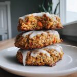 Three Soft-Baked Carrot Cake Cookies stacked on a white plate, drizzled with cream cheese glaze.