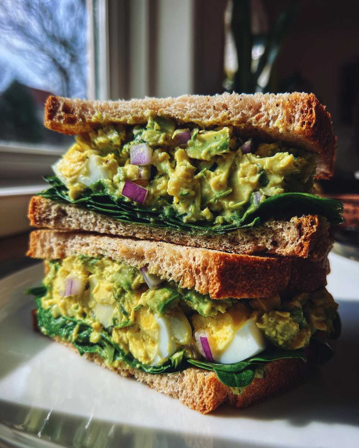 A tall, stacked Avocado Egg Salad Sandwich featuring creamy avocado filling, sliced hard-boiled eggs, red onion, and spinach on whole wheat bread.