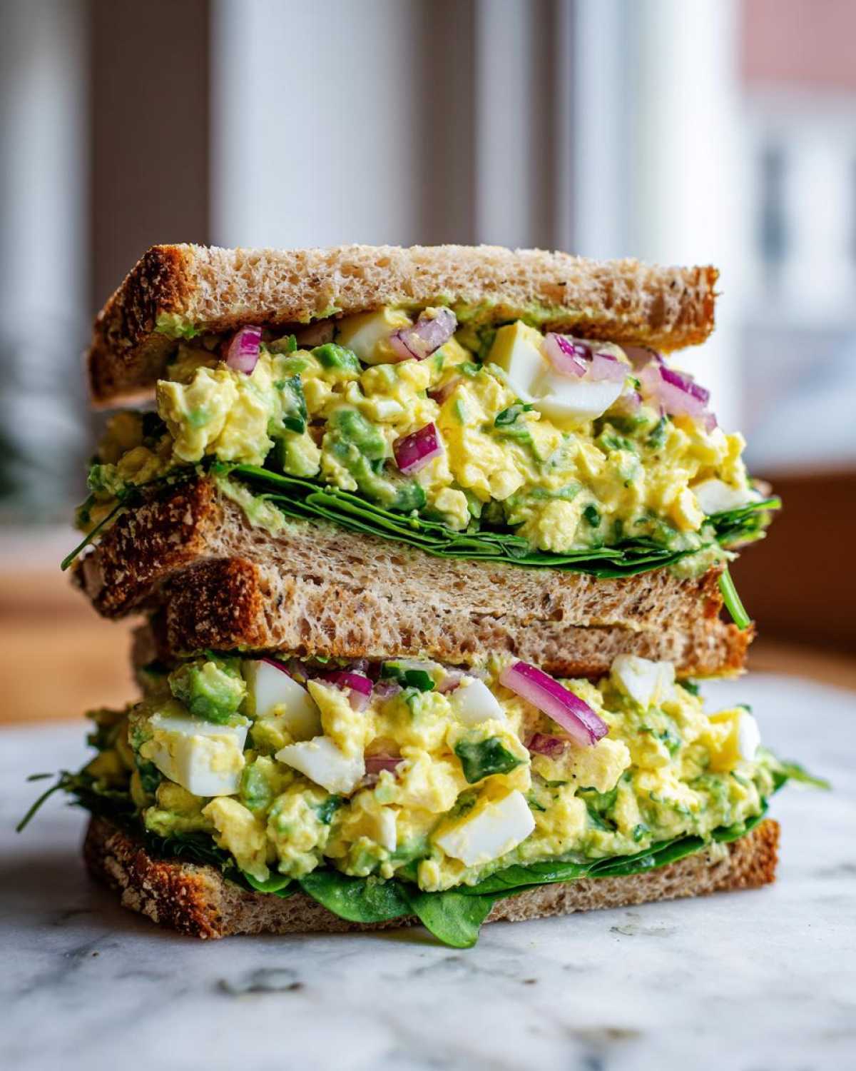 Two halves of a stacked Avocado Egg Salad Sandwich on whole wheat bread, filled with creamy avocado, chopped egg, and red onion.