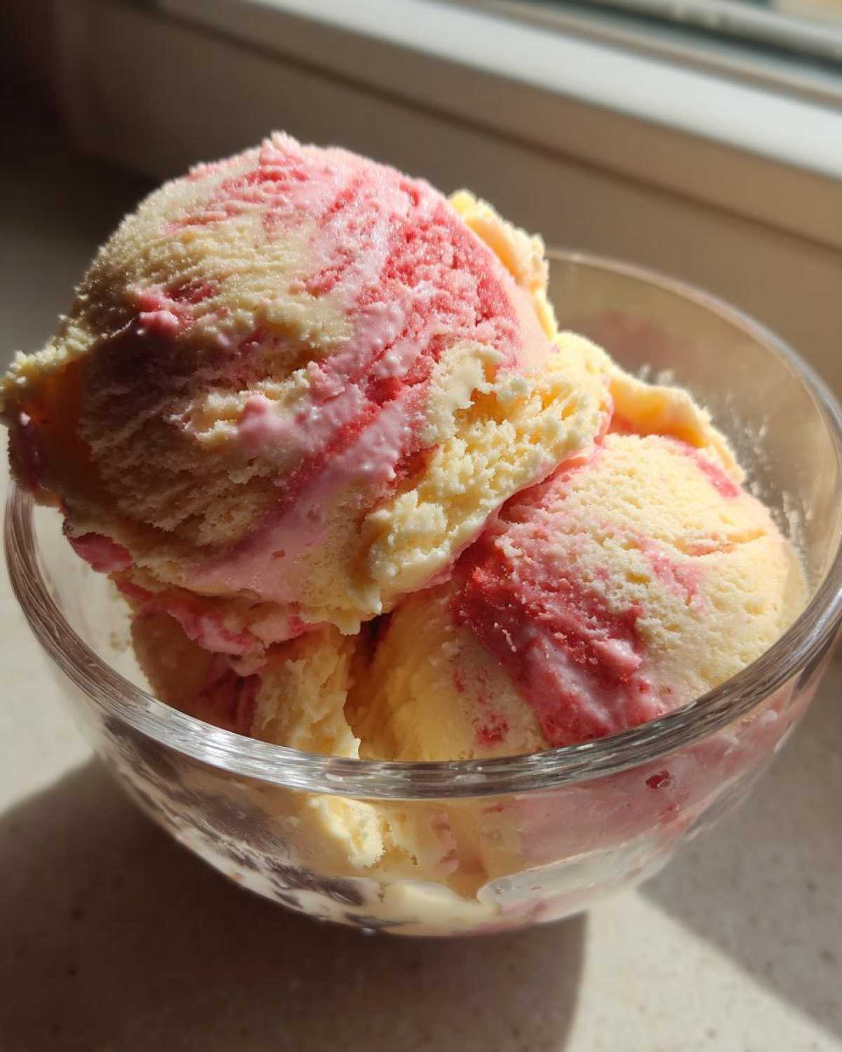 Three scoops of swirled Strawberry Banana Ice Cream, showing yellow and pink streaks, served in a clear glass bowl.