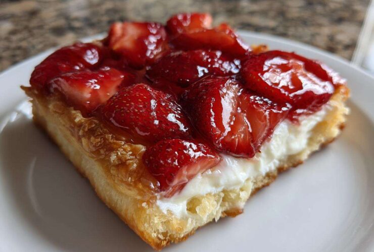 A close-up view of a flaky pastry square topped with cream cheese filling and glazed strawberries, a perfect Strawberry Cream Cheese Danish.