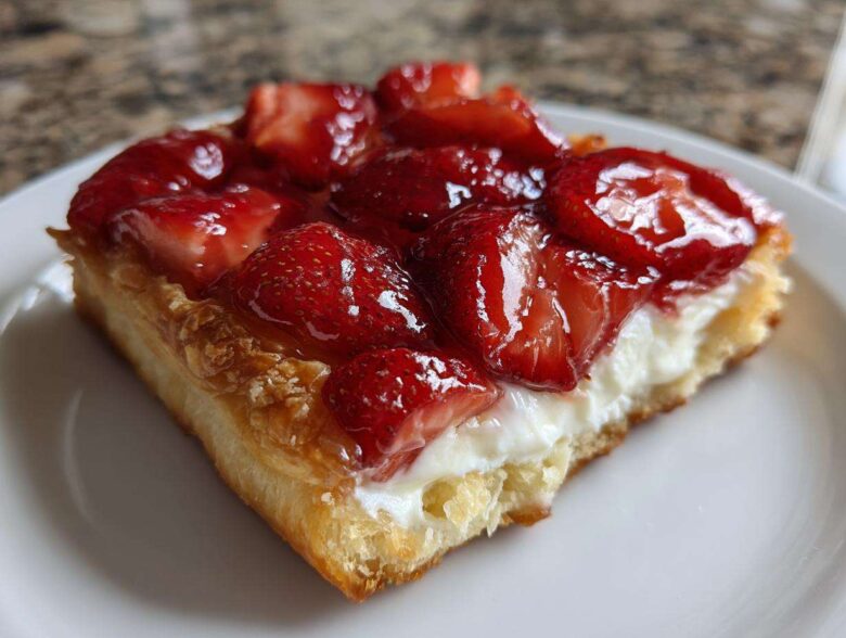 A close-up view of a flaky pastry square topped with cream cheese filling and glazed strawberries, a perfect Strawberry Cream Cheese Danish.