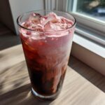 A tall glass of layered Strawberry Cream Cold Brew with ice, sitting on a wooden surface near a window.
