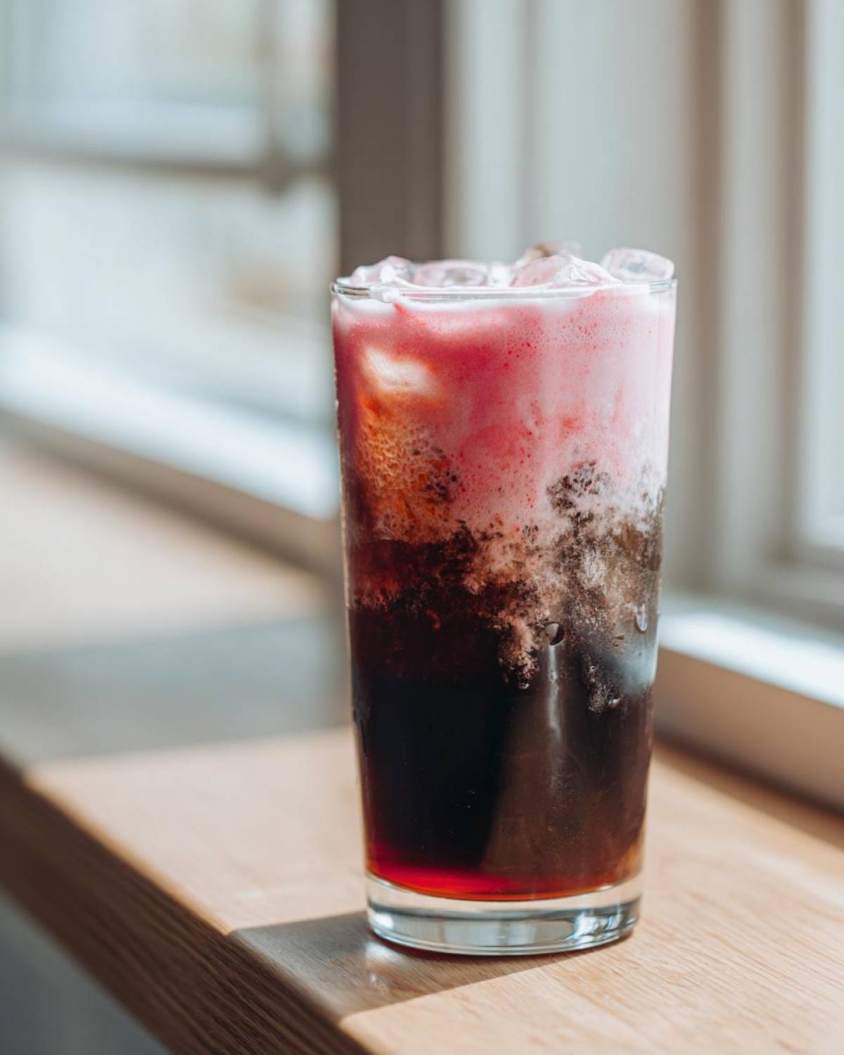 A tall glass of layered Strawberry Cream Cold Brew with ice, sitting on a wooden surface near a window.