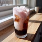 A tall glass of Strawberry Cream Cold Brew with dark coffee and pink foam topping, sitting in bright sunlight.