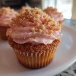 A close-up of a Strawberry Crunch Cupcakes featuring pink strawberry frosting and a generous topping of crunchy golden crumbs.