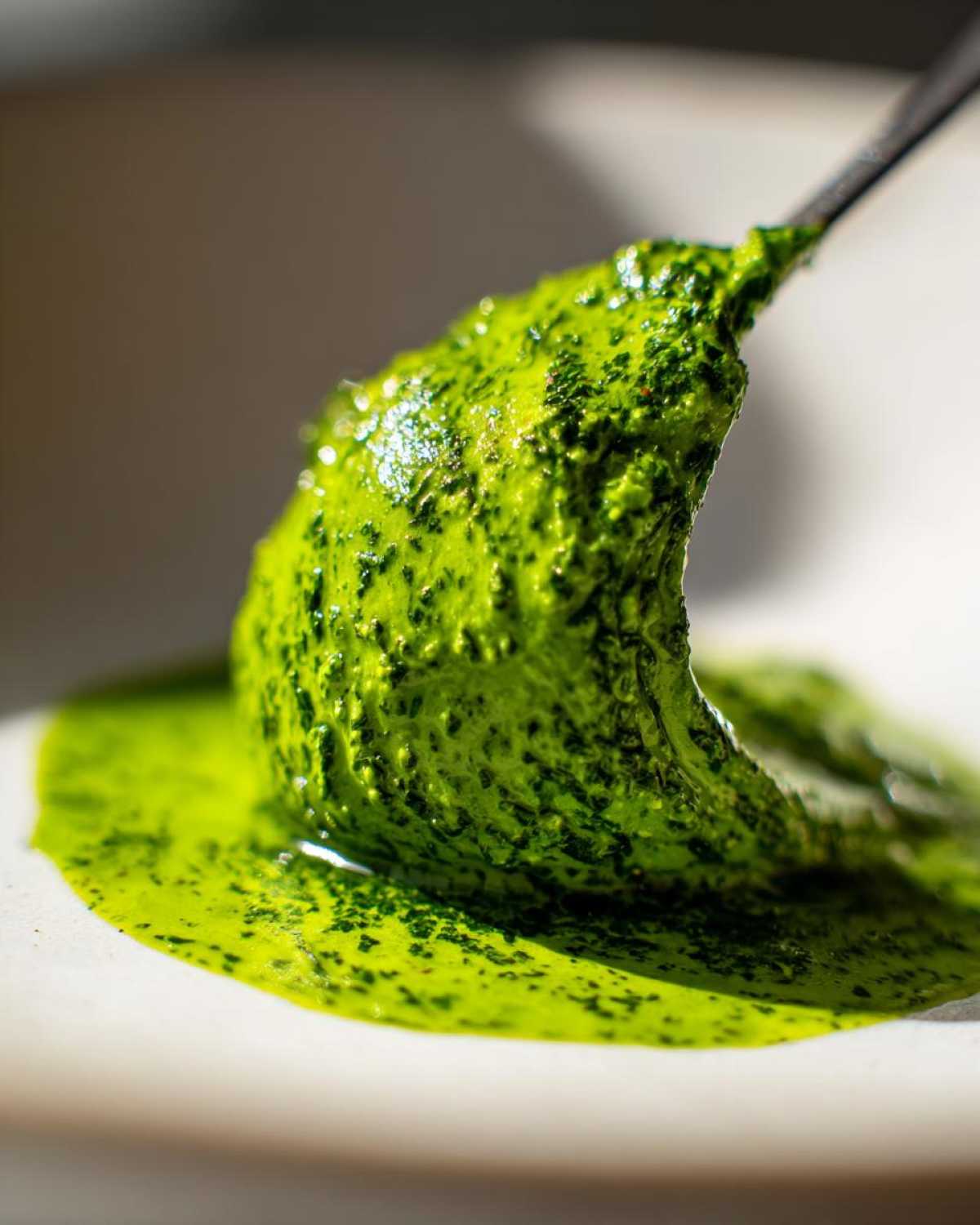 A close-up, macro shot showing a spoonful of bright green Vegan Basil Pesto dripping onto a light surface.
