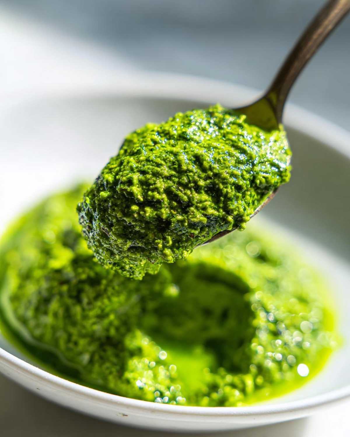 A spoonful of vibrant green Vegan Basil Pesto being lifted from a white bowl, showing its thick, textured consistency.