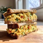 Two halves of a Vegan Chickpea Salad Sandwich stacked on a wooden board, showing creamy yellow filling with visible chickpeas and vegetables.