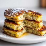 Close-up of a stack of fluffy Vegan Lemon Poppy Seed Pancakes dusted with powdered sugar and drizzled with syrup.