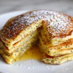 A stack of fluffy Vegan Lemon Poppy Seed Pancakes, cut to show the interior, drizzled with syrup and dusted with powdered sugar.