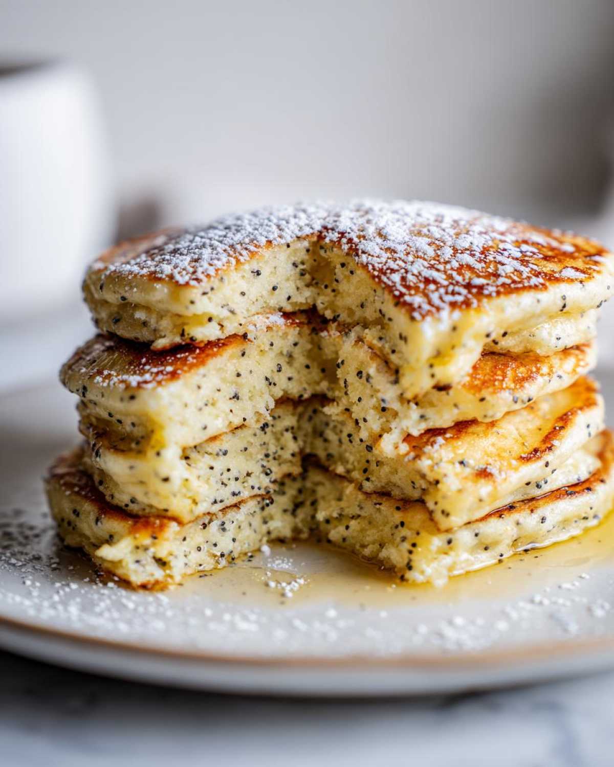 Close-up of a stack of fluffy Vegan Lemon Poppy Seed Pancakes dusted with powdered sugar and drizzled with syrup.