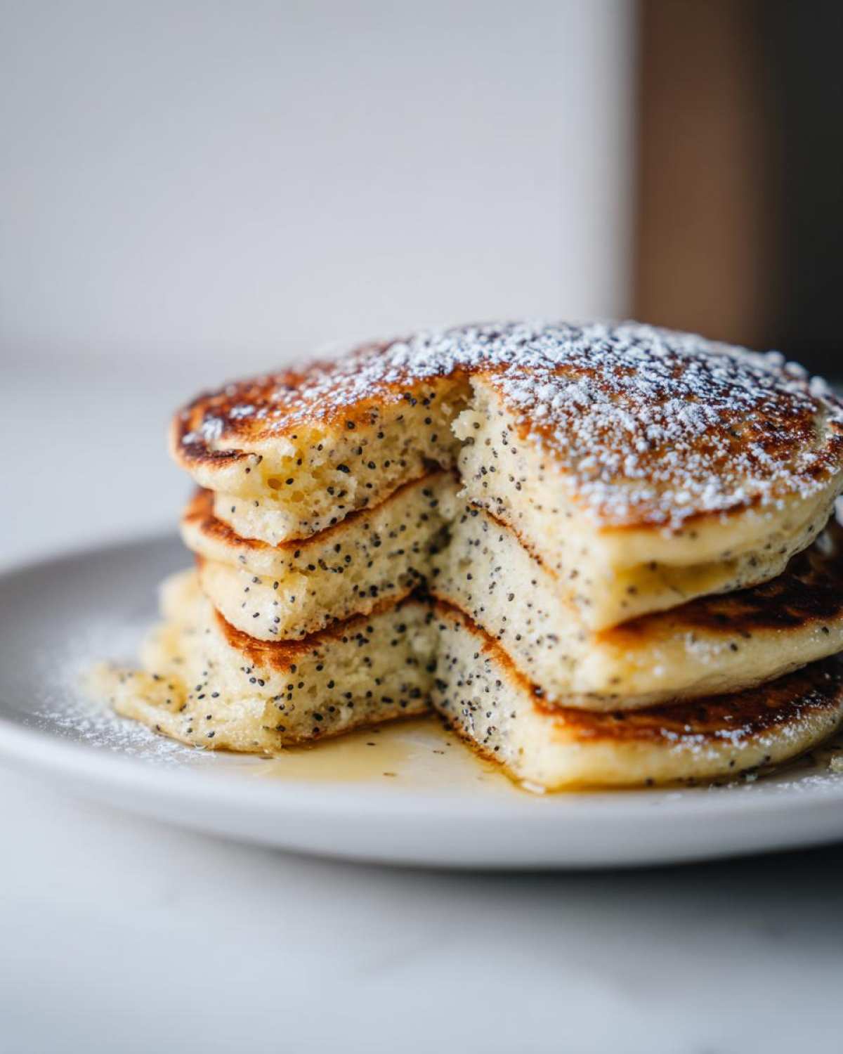 A stack of fluffy Vegan Lemon Poppy Seed Pancakes dusted with powdered sugar and drizzled with syrup.