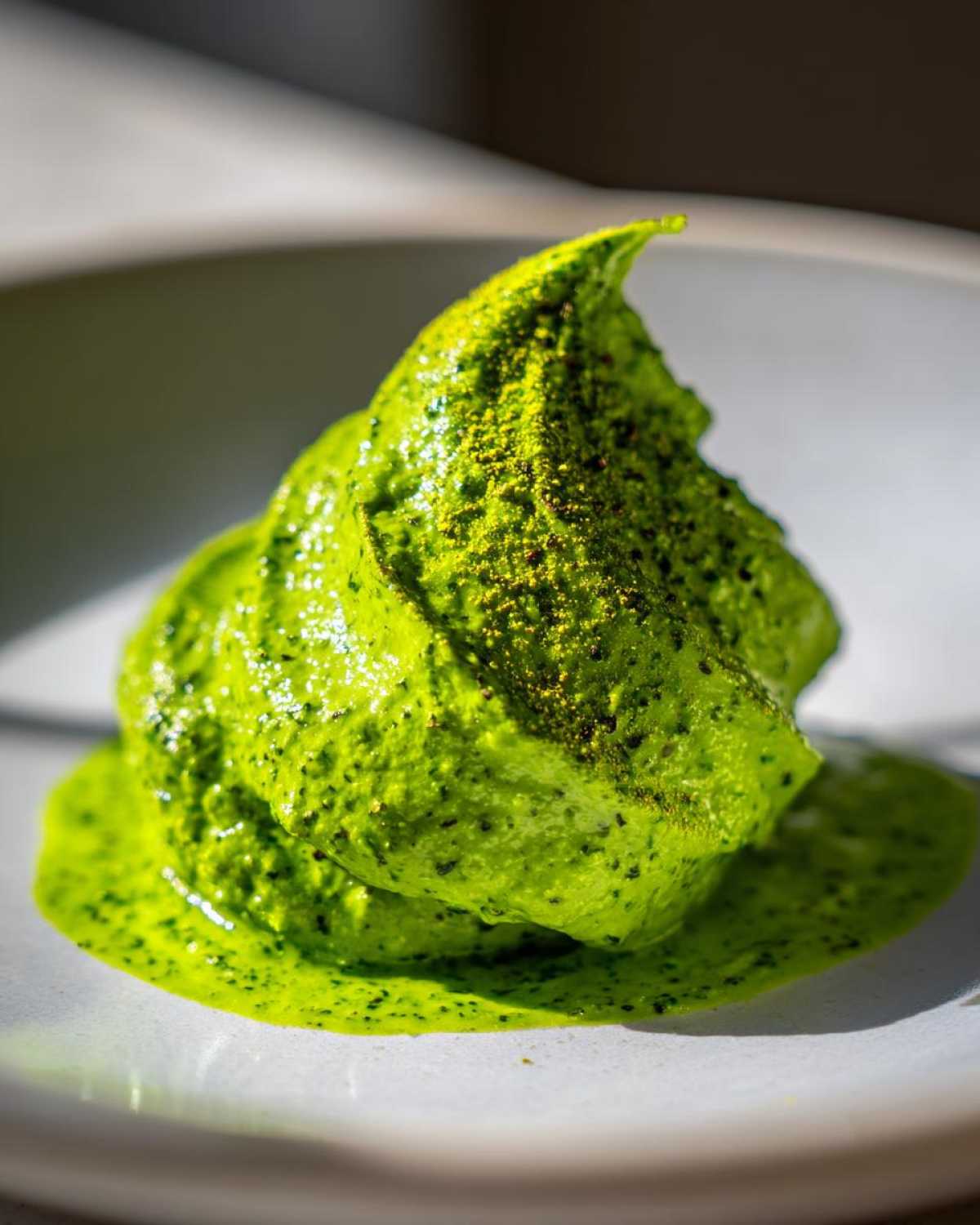 Close-up of a bright green, thick scoop of Vegan Basil Pesto, dusted with fine seasoning, on a light plate.