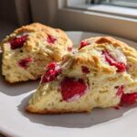 Two freshly baked White Chocolate Raspberry Scones, one sliced open showing raspberries and white chocolate drizzle, on a white plate.