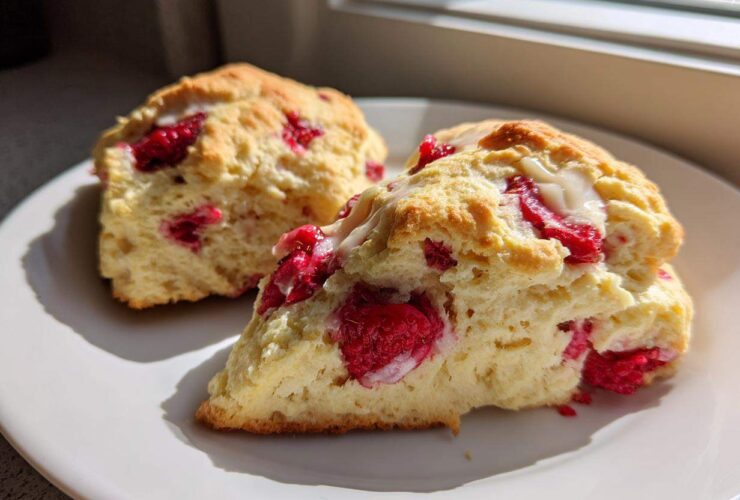 Two freshly baked White Chocolate Raspberry Scones, one sliced open showing raspberries and white chocolate drizzle, on a white plate.
