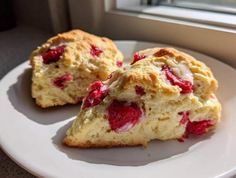 Two freshly baked White Chocolate Raspberry Scones, one sliced open showing raspberries and white chocolate drizzle, on a white plate.