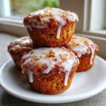 Three golden brown Apple Fritter Muffins topped with a thick, dripping vanilla glaze, stacked on a white plate.