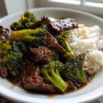 A close-up of a white bowl filled with Beef and Broccoli Stir Fry served next to fluffy white rice.