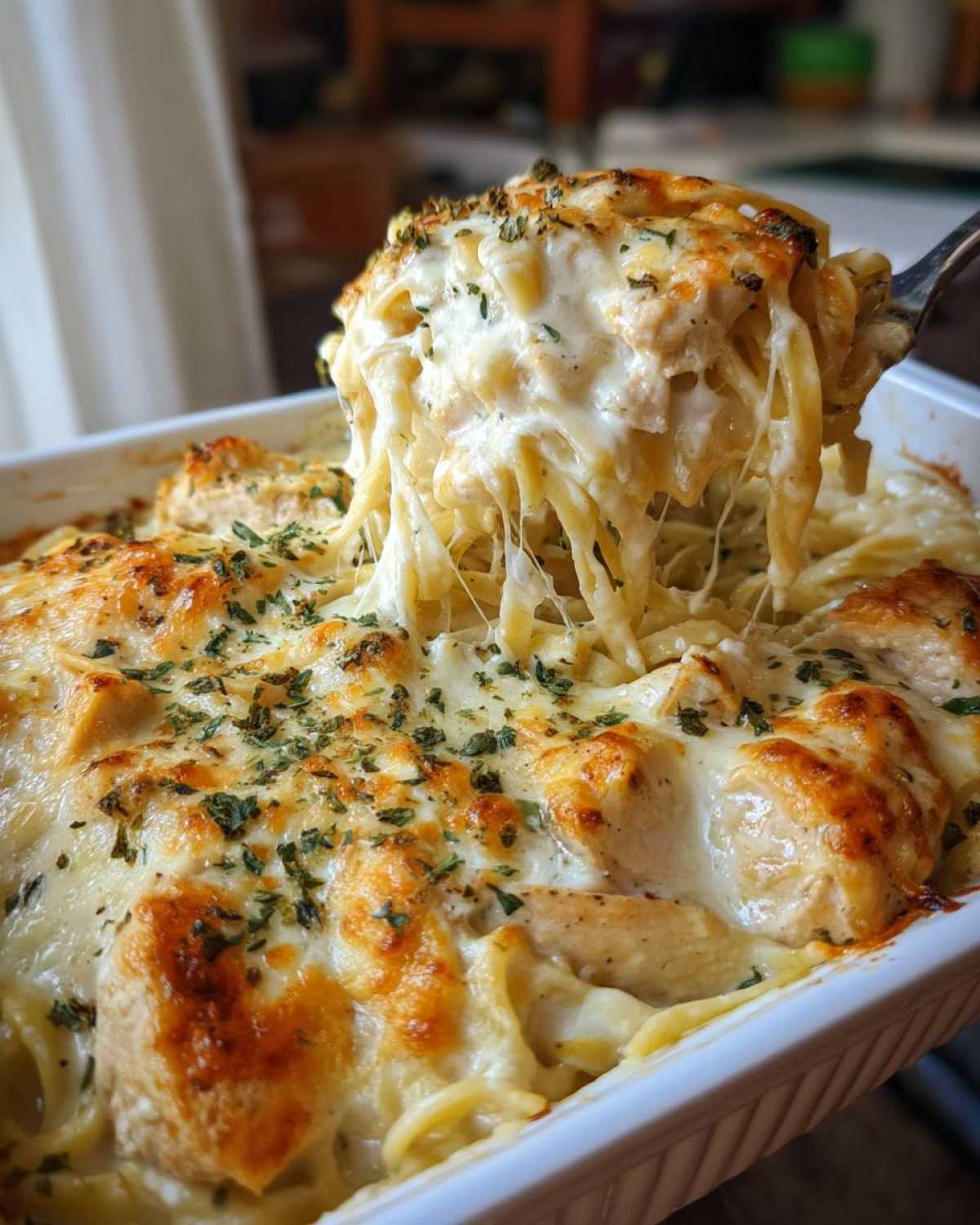 A fork lifts a serving of Cheesy Chicken Alfredo Bake, showing long, stretchy strands of melted cheese.