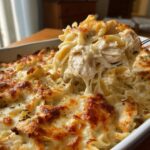 A fork lifts a serving of Cheesy Chicken Alfredo Bake, showing shredded chicken and pasta coated in creamy sauce and topped with browned cheese.
