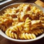 Close-up of fusilli pasta coated in a rich, creamy, cheesy sauce with chunks of chicken in a white bowl.