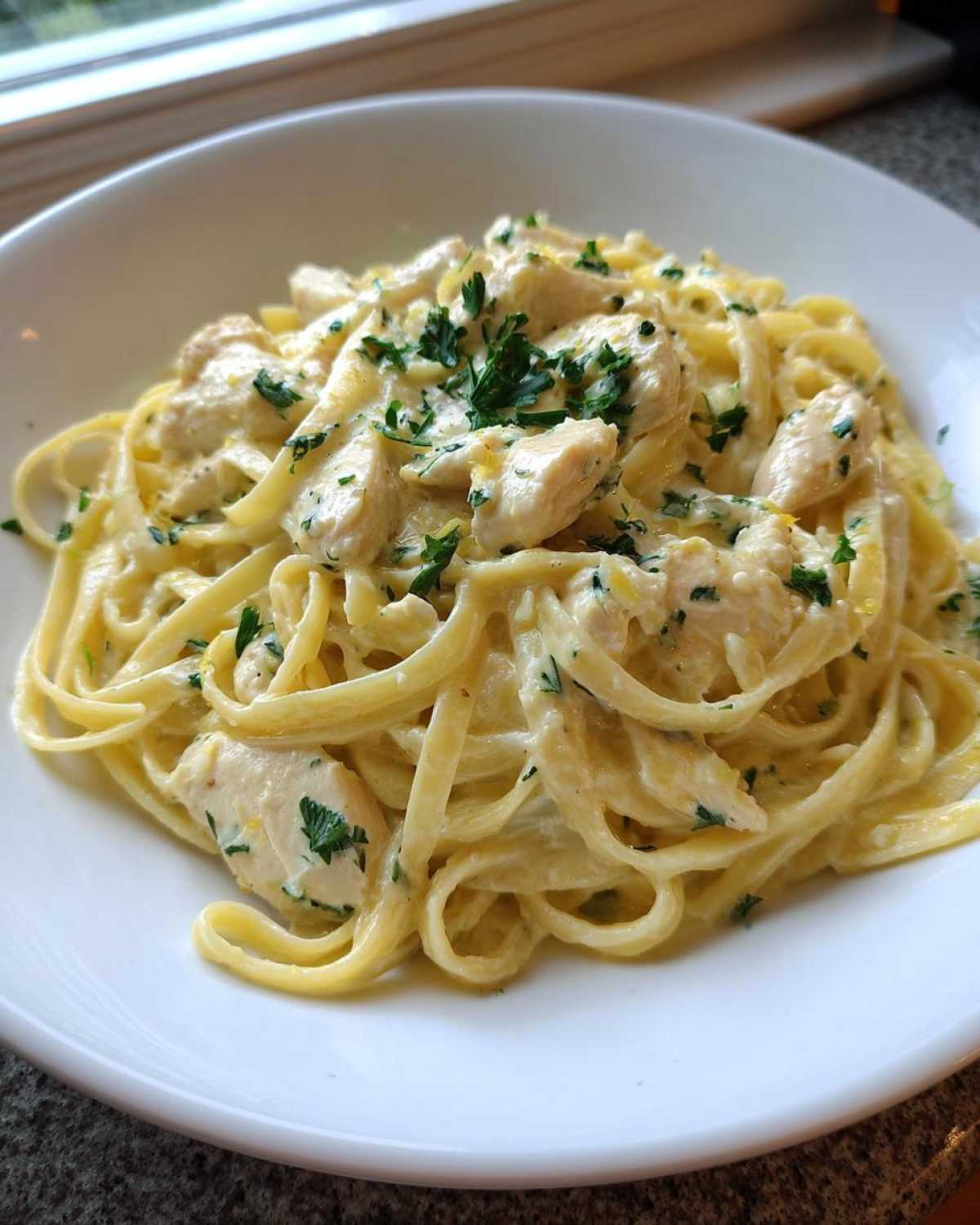 A close-up of fettuccine noodles coated in a rich sauce with chunks of chicken, garnished with parsley, in Creamy Lemon Garlic Pasta with Chicken.
