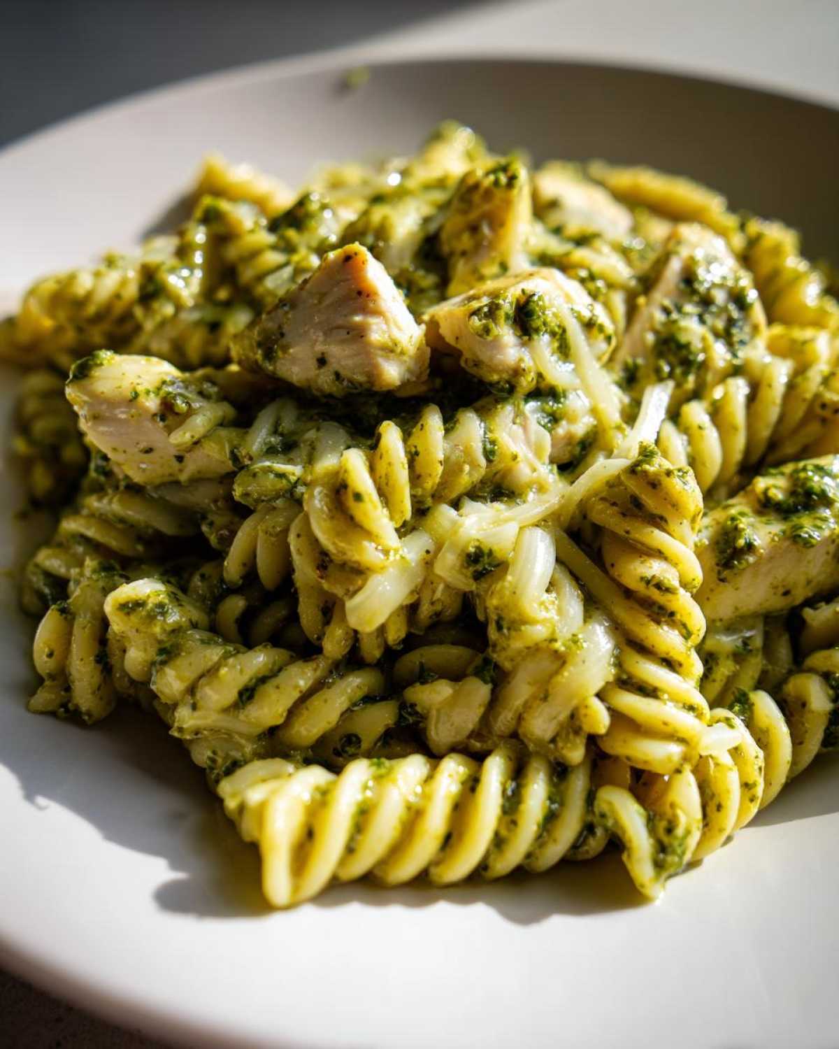 Close-up shot of creamy pesto chicken pasta with fusilli noodles, chunks of chicken, and melted mozzarella cheese.