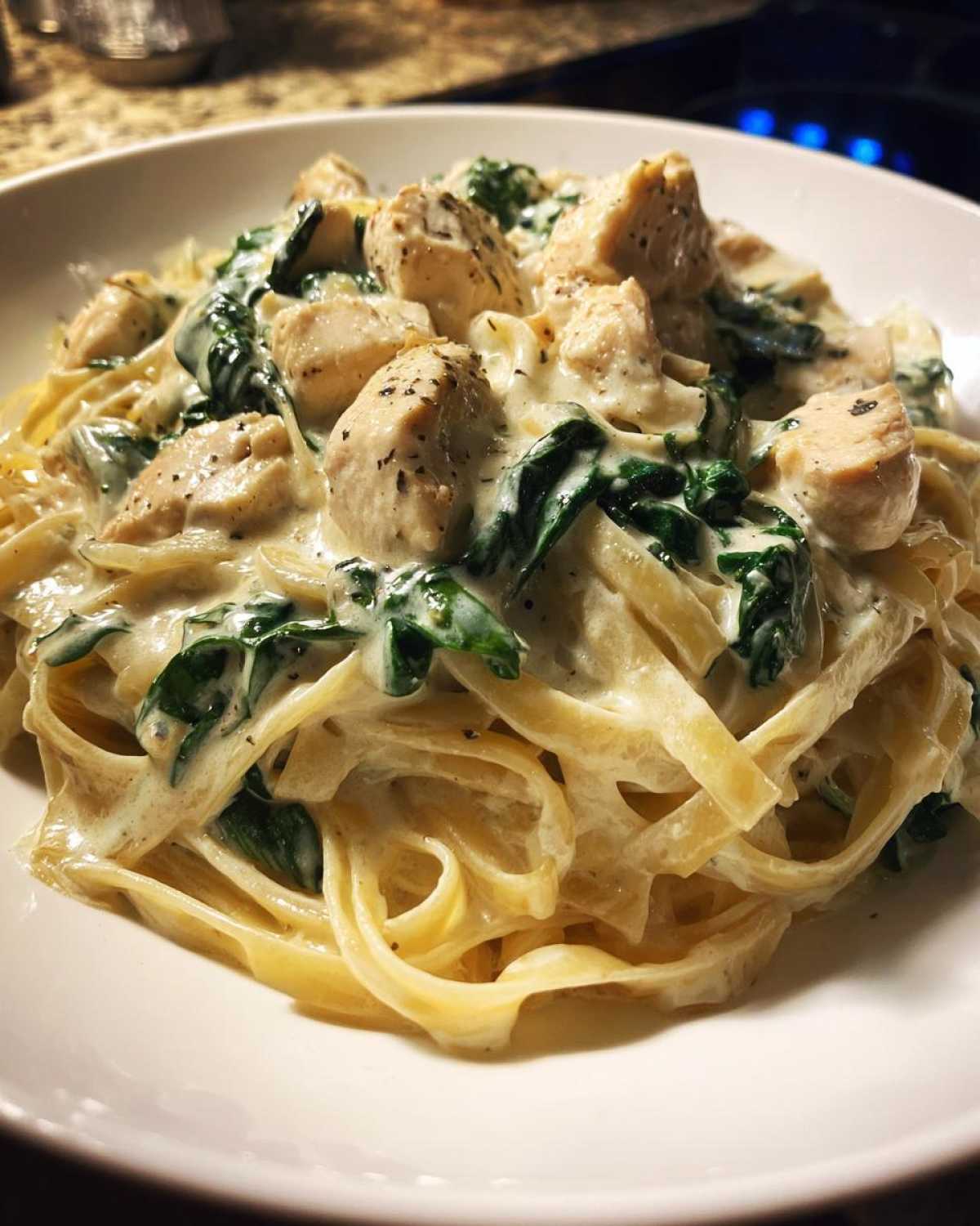 A close-up of a white bowl filled with creamy spinach pasta with chicken dinner, featuring fettuccine noodles and seasoned chicken pieces.