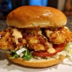 A close-up of a Crispy Chicken Burger with Honey Mustard Sauce, featuring a golden bun, fried chicken, lettuce, tomato, and sauce.