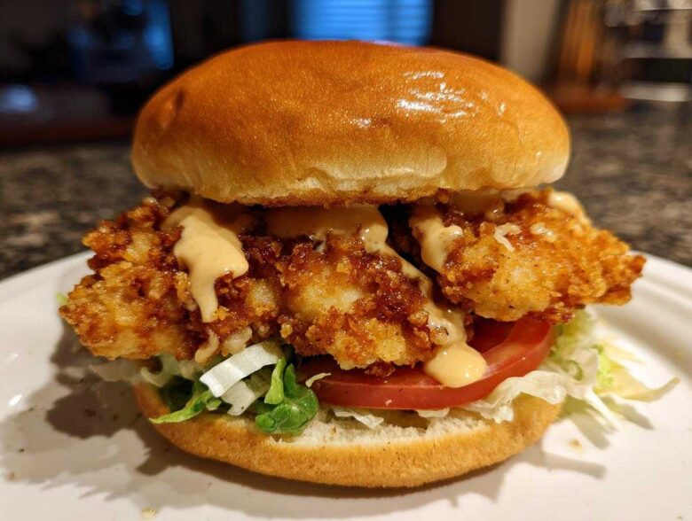 A close-up of a Crispy Chicken Burger with Honey Mustard Sauce, featuring a golden bun, fried chicken, lettuce, tomato, and sauce.