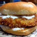 A close-up of a Crispy Chicken Sandwich with Garlic Sauce featuring a golden, crunchy fried chicken fillet between a toasted bun, drizzled with white sauce.
