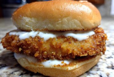 A close-up of a Crispy Chicken Sandwich with Garlic Sauce featuring a golden, crunchy fried chicken fillet between a toasted bun, drizzled with white sauce.