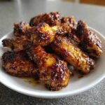 A close-up of several crispy Honey Garlic Chicken Wings coated in a sticky, dark glaze, piled on a white plate.