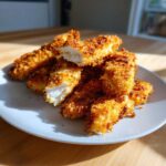 A pile of golden brown Crispy Oven Baked Chicken Tenders Extra Juicy, with one piece cut open showing the moist white interior.