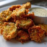 A close-up of golden brown Delicious Air Fryer Fried Pickles stacked on a white plate next to a small bowl of dipping sauce.