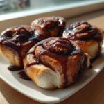Four freshly baked coffee rolls generously drizzled with dark, shiny glaze, served on a white plate.