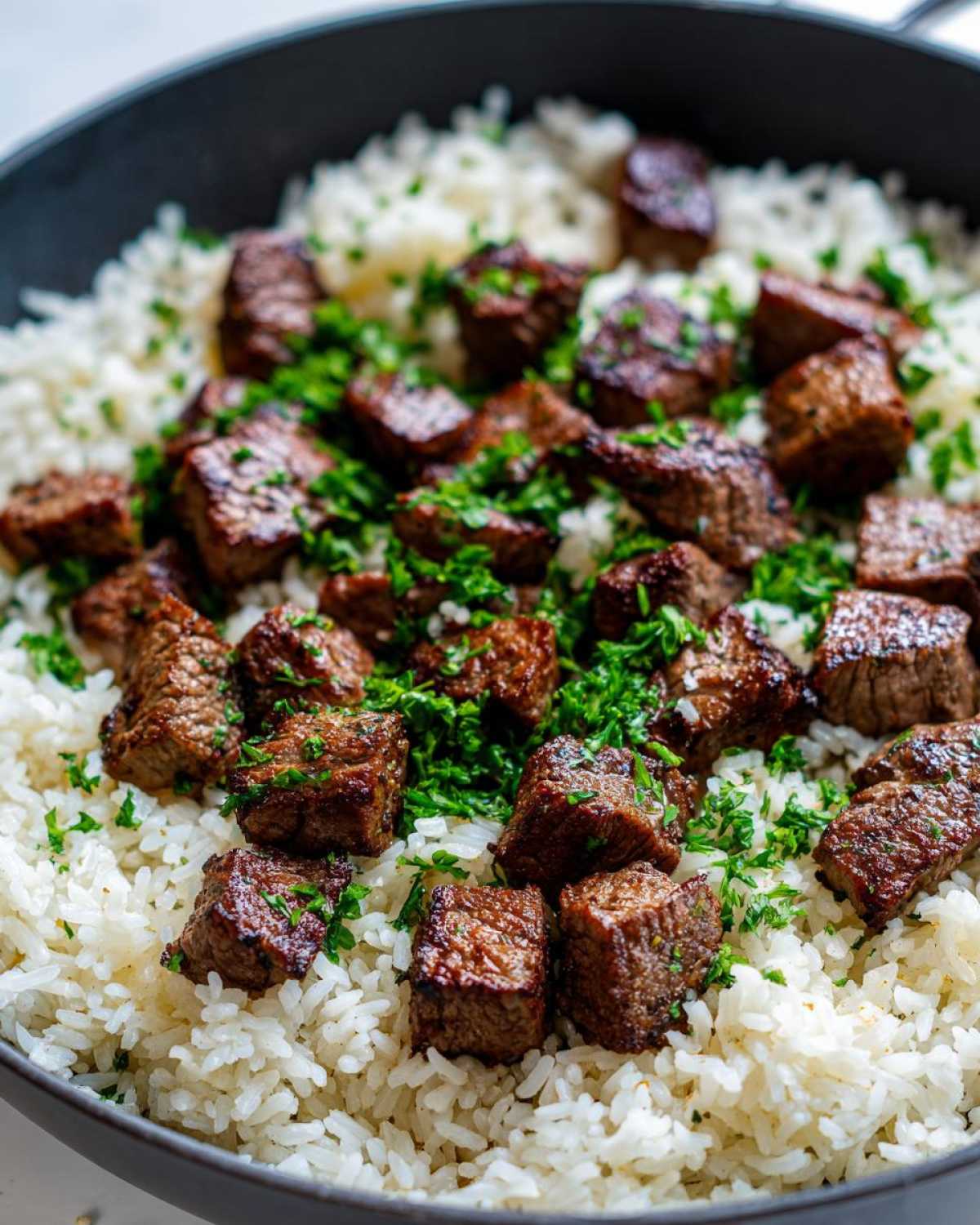 Close-up of seared steak bites served over fluffy white rice, garnished with fresh parsley in a dark skillet, showcasing the Garlic Butter Steak and Rice Skillet Dinner.