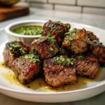Close-up of seared Garlic Butter Steak Bites topped with green herb sauce, served on a white plate.
