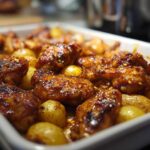 Close-up of glossy, baked chicken pieces coated in honey mustard glaze nestled among roasted potatoes in a white baking dish.