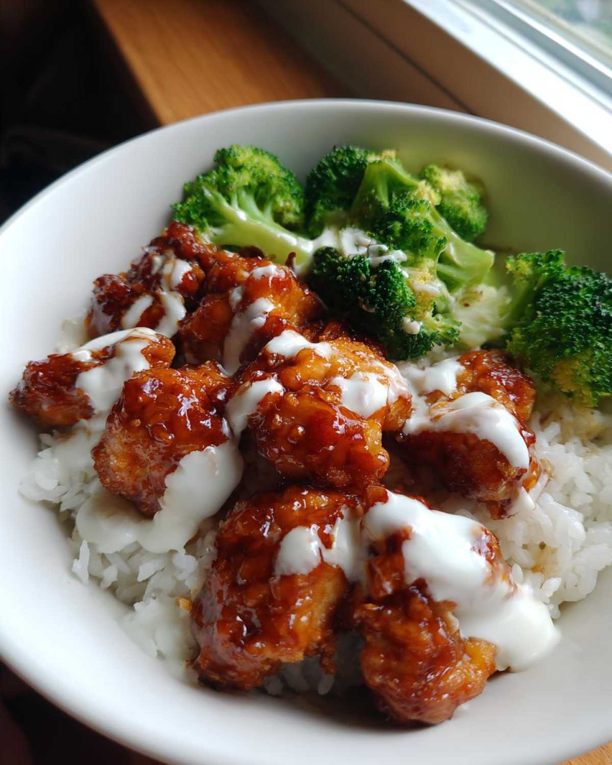 Close-up of a Honey Soy Chicken Bowl with sticky glazed chicken pieces, white rice, broccoli, and a drizzle of white garlic sauce.