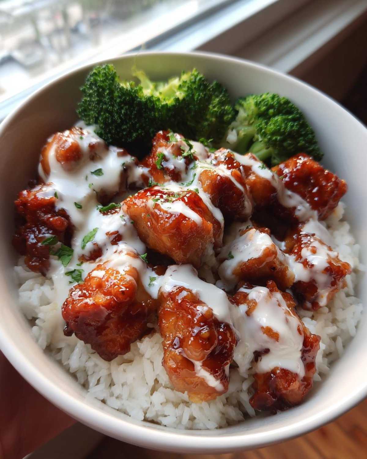 Close-up of a Honey Soy Chicken Bowl with garlic sauce drizzled over glazed chicken pieces served on white rice with steamed broccoli.