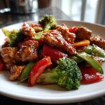 Close-up of Honey Soy Chicken Skillet with Veggies featuring glossy chicken pieces, red peppers, broccoli, and snow peas on a white plate.